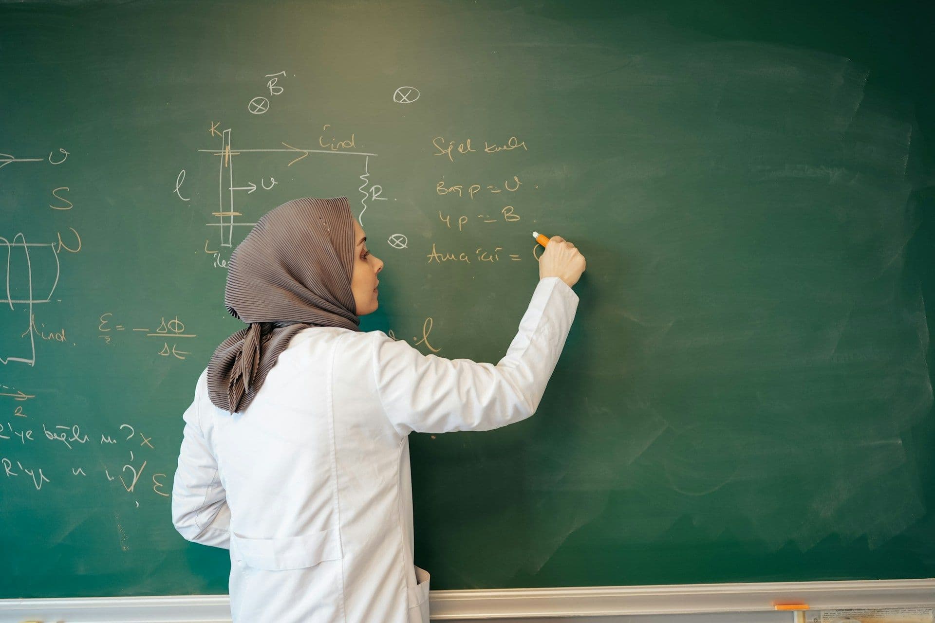A teacher in a headscarf writing on a chalkboard in front of her class
